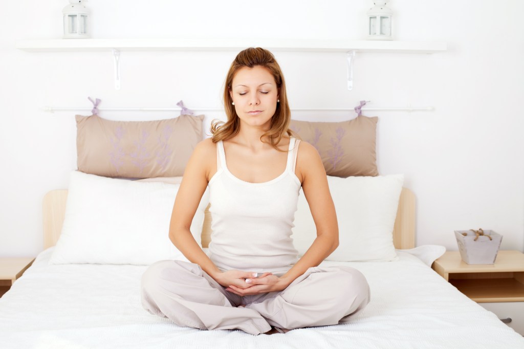 Woman in white sitting on a bed in meditating pose Como lidar com a ansiedade do isolamento social?